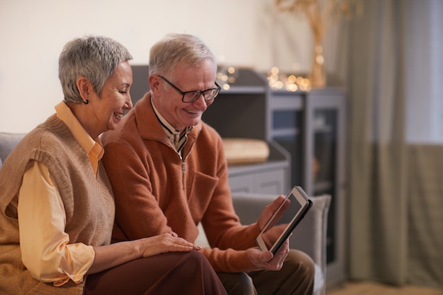 An elderly couple sit on a grey couch in their rental unit and enjoy a video tour of a rental property